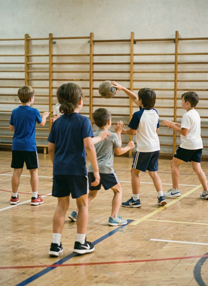 Groep kinderen van een jaar of negen in een gymzaal die met een kleine handbal oefent op passen en vangen, begeleider op de achtergrond